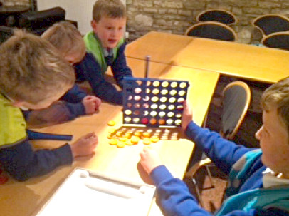Focused children engage each other's wits in a battle of Connect 4, whilst others look on learning from their peers, offering advice, engaging in conversation.
